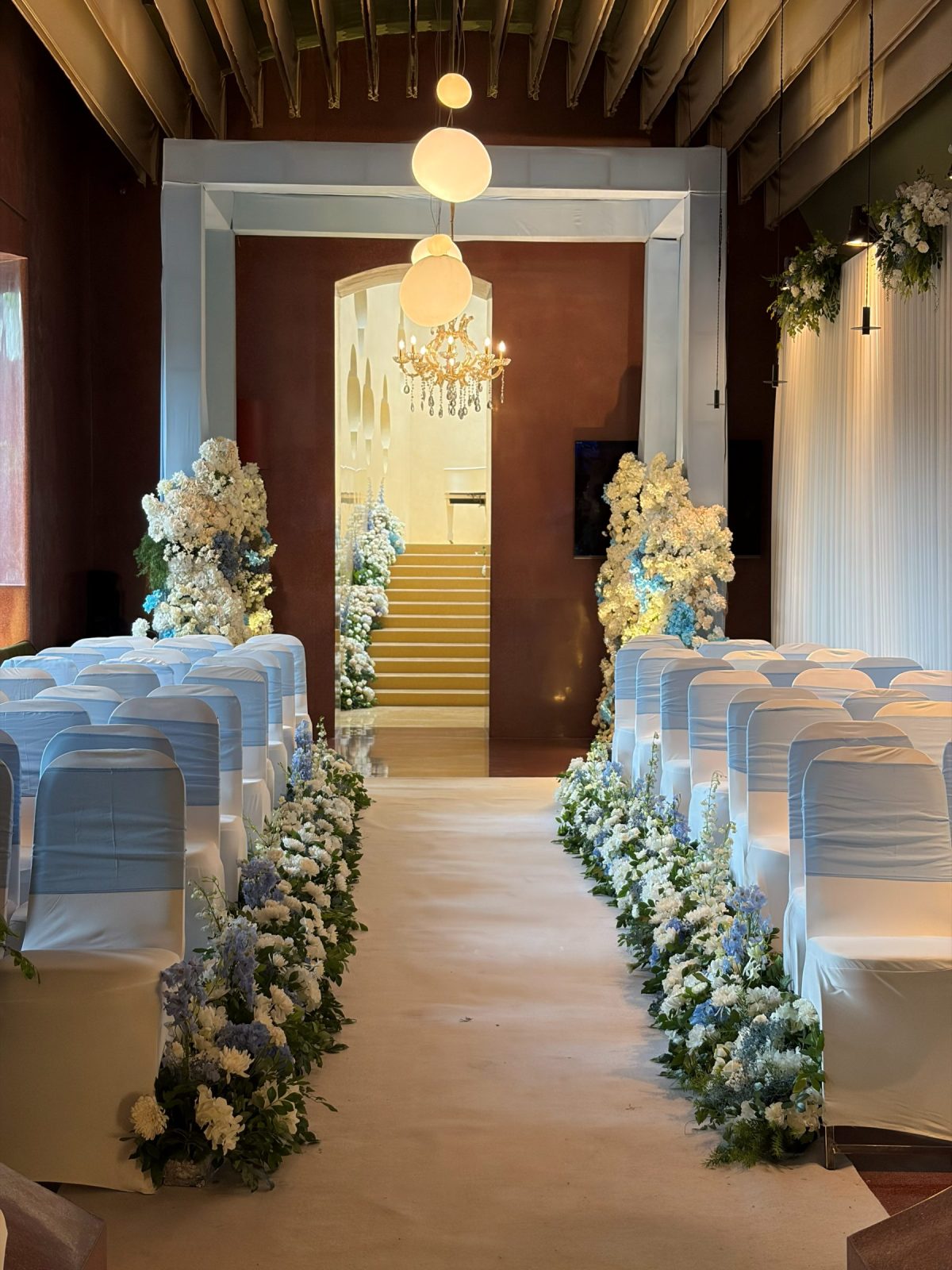 Ceremony aisle lined with white and powder-blue florals, chairs dressed in ivory covers