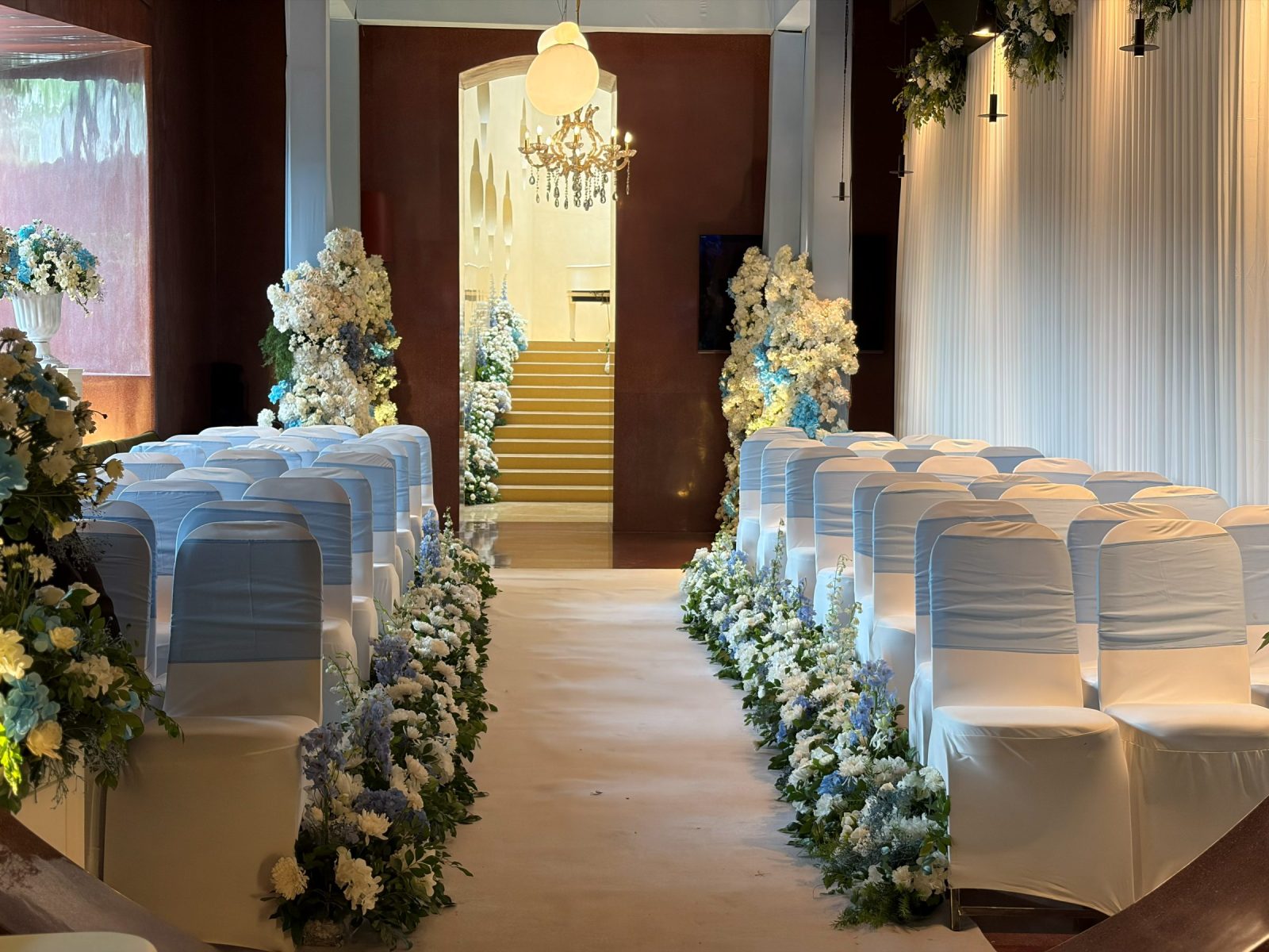 Ceremony aisle lined with white and powder-blue florals, chairs dressed in ivory covers